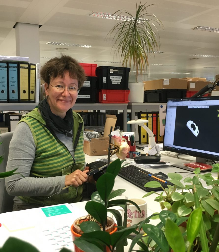 Designer Petra at her workplace. She sits at a corner desk. In addition to the PC, keyboard and other office supplies, there are also various plants on her table. In the background there is a shelf with many folders.