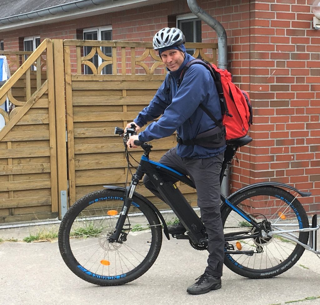 Equipped with weatherproof clothing, a helmet and a red backpack, Johannes sits on his bike. A house can be seen behind him.