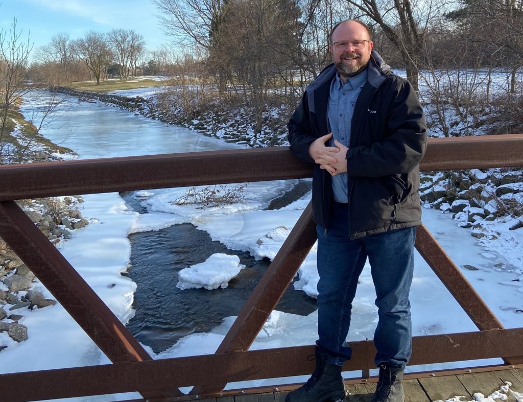 Richard is leaning on a bridge that leads over a partially frozen river. The trees are bare, there is some snow and the sun is shining.