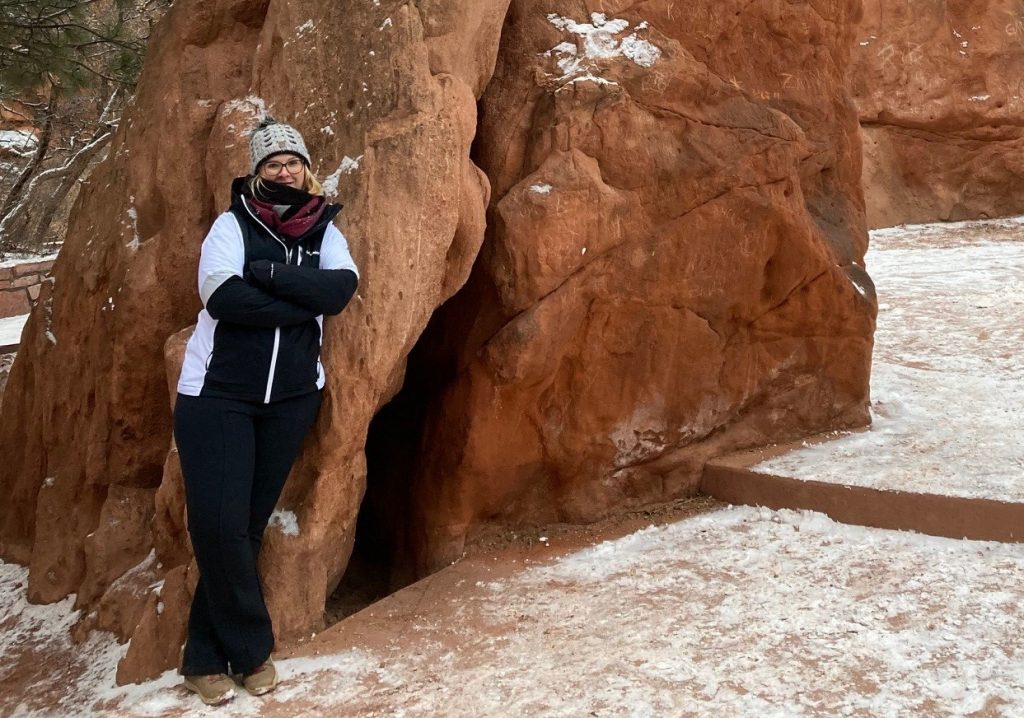 Nadine leans against a large rock. She wears thick winter clothes, gloves, scarf and hat. There is light snow on the ground.