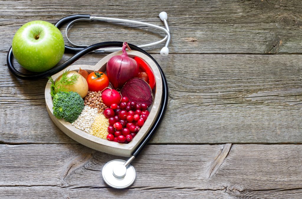 A heart-shaped bowl lies on a wooden floor. There are different types of vegetables in it. Next to it lies a green apple and a stetoscope.