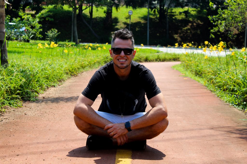 Deniz sits cross-legged on the ground in the sun in the park and smiles at the camera.