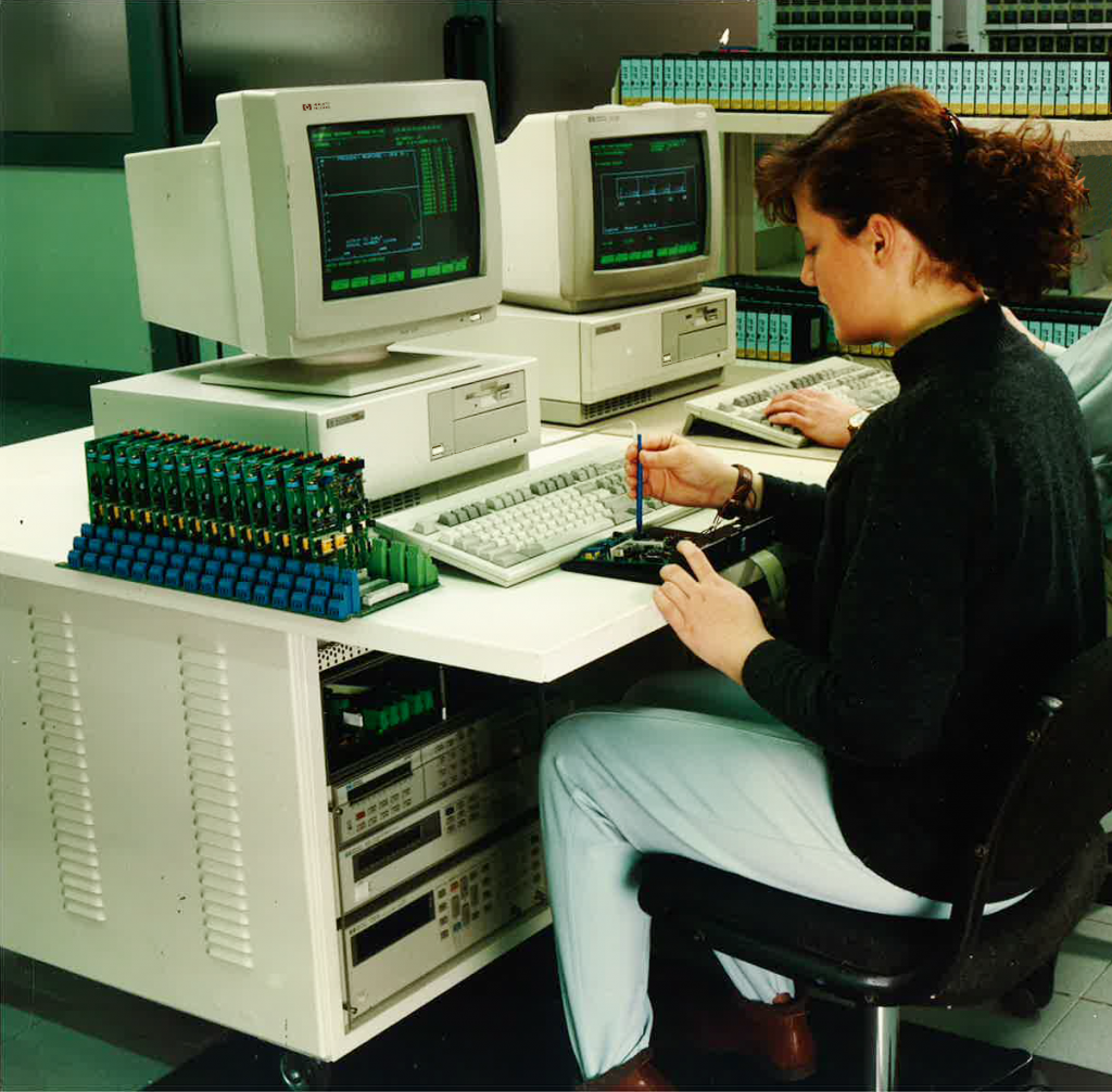 In 1994, Marta Bariselli is seated at her workstation in production planning. On the table in front of her is a PC with a cathode-ray tube monitor. She is currently working on a small circuit board placed directly in front of her.