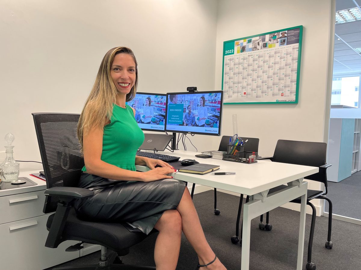 Carina, Regional HR Director Latam, sits in an office chair in her office, facing the camera and smiling. Behind her on the desk, you can see two monitors, pens, and a notebook.