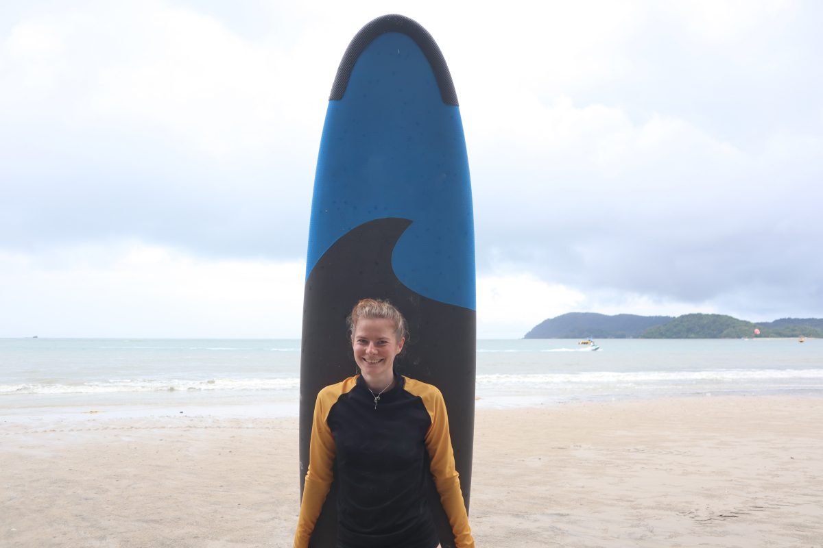 Trainee Clara stands on the beach with her surfboard.