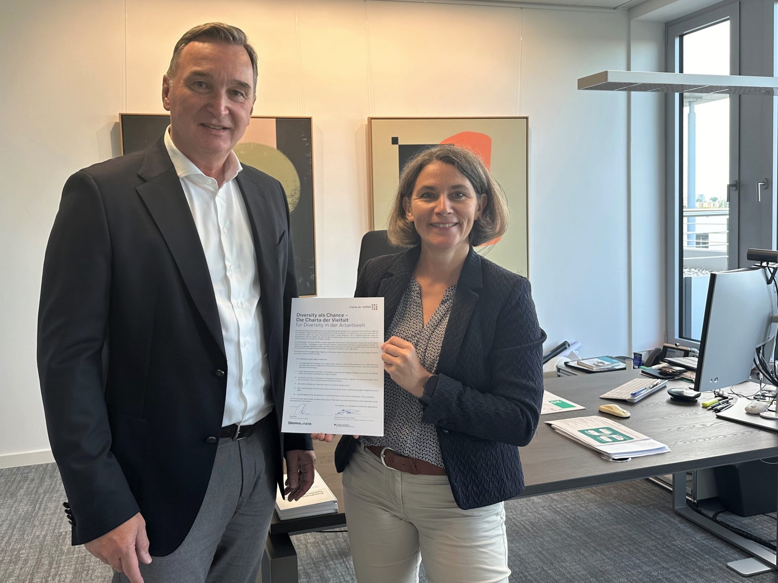 Pepperl+Fuchs CHRO Florian Ochs and Cordula Straub, founding member of the Diversity+Inclusion team, stand next to each other facing the camera. xxx holds the signed document of the Diversity Charter up to the camera.