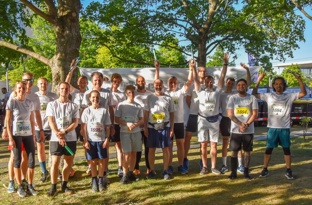 Group photo of the Pepperl+Fuchs runners at the relay race in Berlin. They are standing on a green meadow and some trees can be seen behind them. Three of them raise the relays. They wear the white Pepperl+Fuchs running shirts.