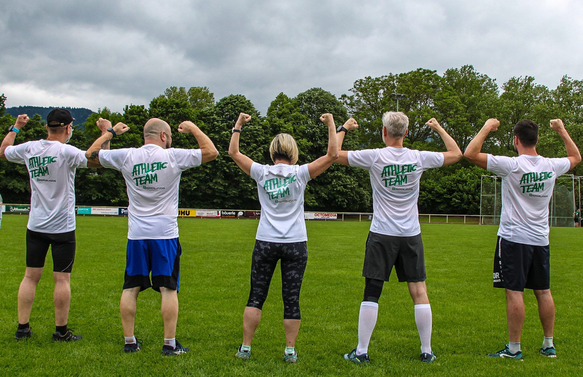 Die Pepperl+Fuchs Läufer*innen beim Firmenlauf in Bühl. Sie stehen auf dem Sportplatz mit dem Rücken zur Kamera und recken ihre Fäuste nach oben. Auf den weißen T-Shirts steht "Athletic Team".