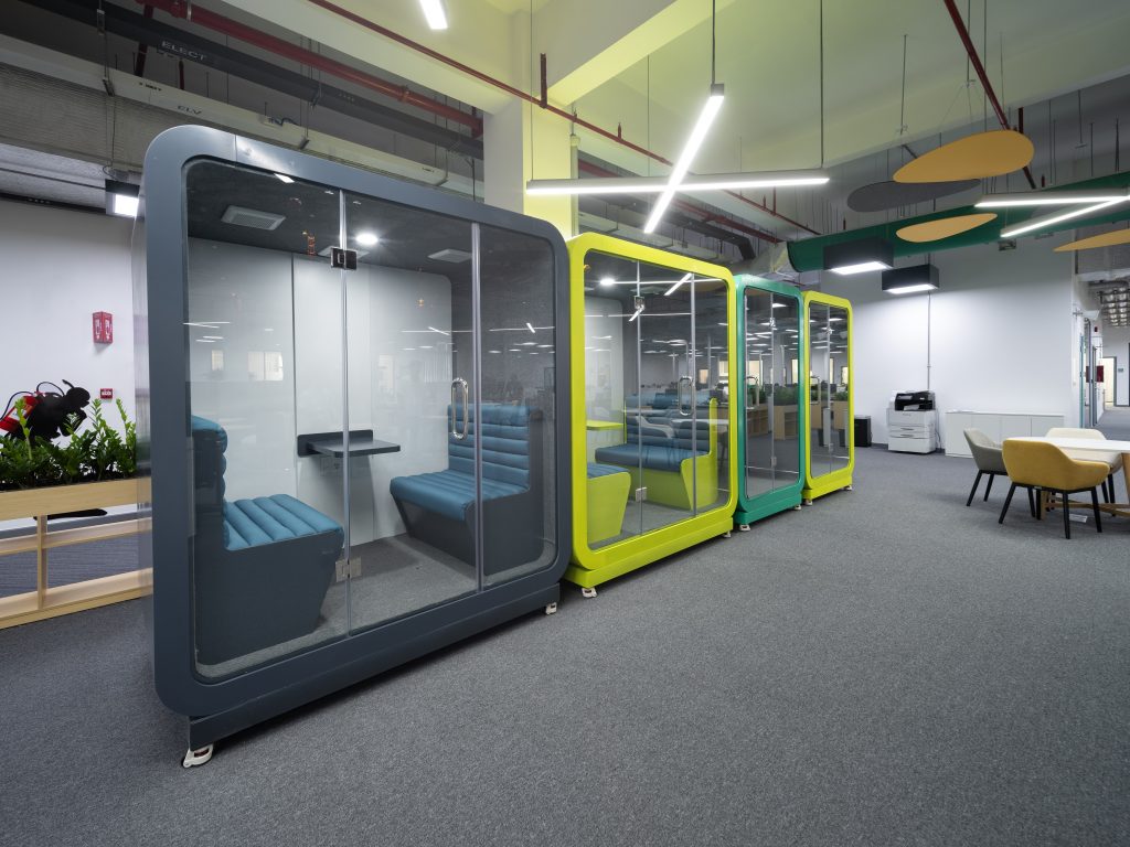 View of four cubes standing in the open-plan office. They are gray, yellow and green and each has a glass door. In a cube there are two small sofas and a small table mounted on the wall.