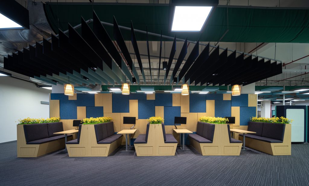 View of four seating niches against a wall. These consist of longer tables with wooden benches with gray upholstery. Yellow flowers are planted between each table.