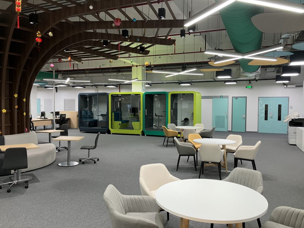 View into the open-plan office. On the left you can see a wooden tree with tables around it. Otherwise there are three more round tables with chairs. In the background you can see four small cubes in which you can have undisturbed conversations.