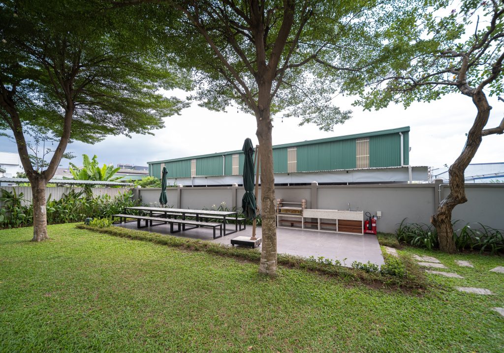 View of the terrace surrounded by trees and meadow. There is a long table with a bench, two parasols and an outdoor kitchen.