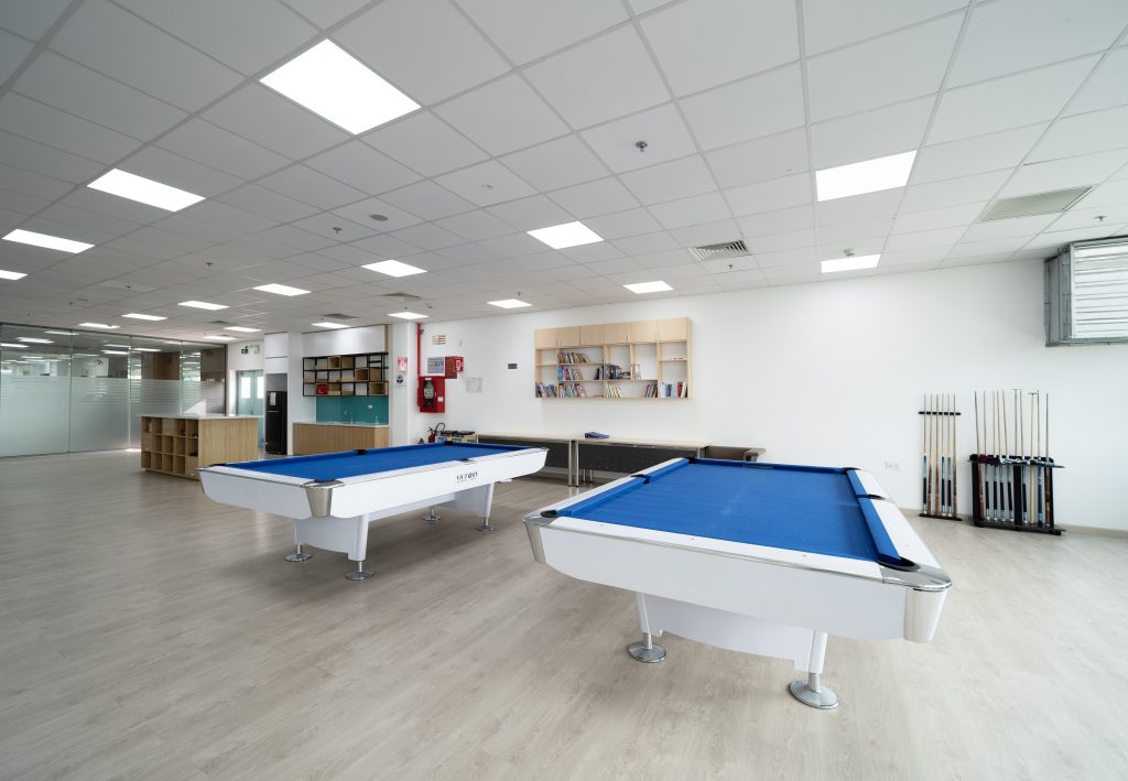 View of two white and blue billiard tables. The reading corner can be seen at the back. There are various books to choose from on the shelves on the wall.