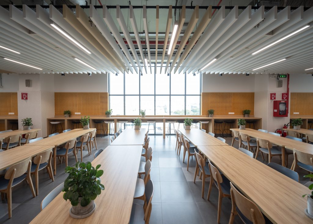 Insight into the canteen at Pepperl+Fuchs Vietnam: view of several rows of tables. The tables are made of light wood and the chairs have white backs. There are small, green plants on the tables.