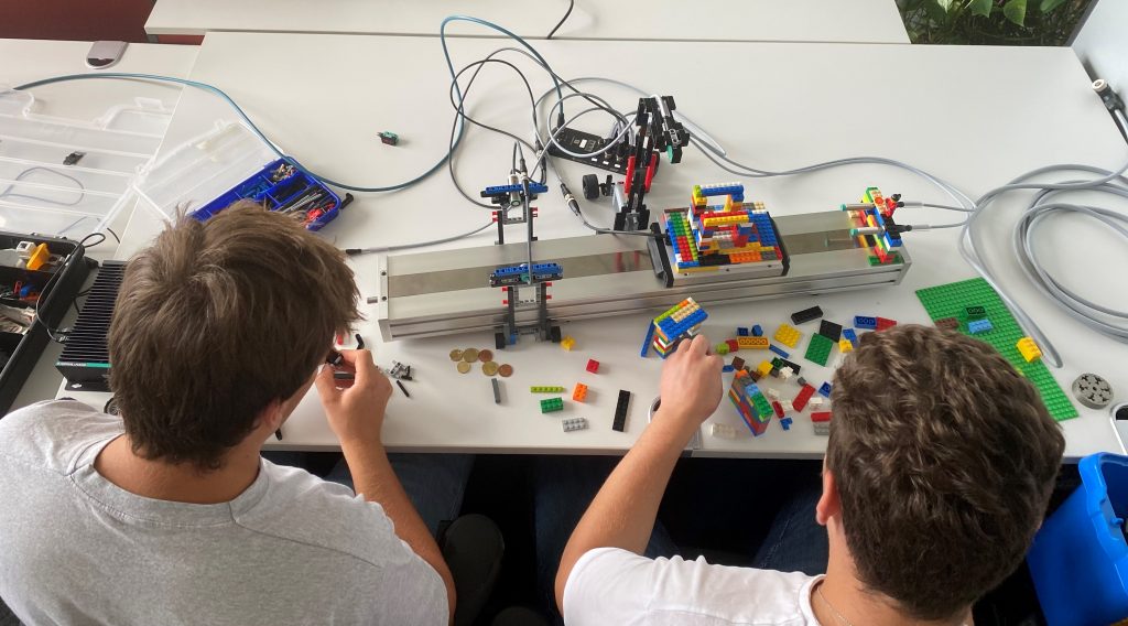 Looking over the shoulder of Aaron and Fabian, who are both studying electrical engineering at Pepperl+Fuchs. They are currently building the holder with Lego blocks. The previous construction is in front of you.