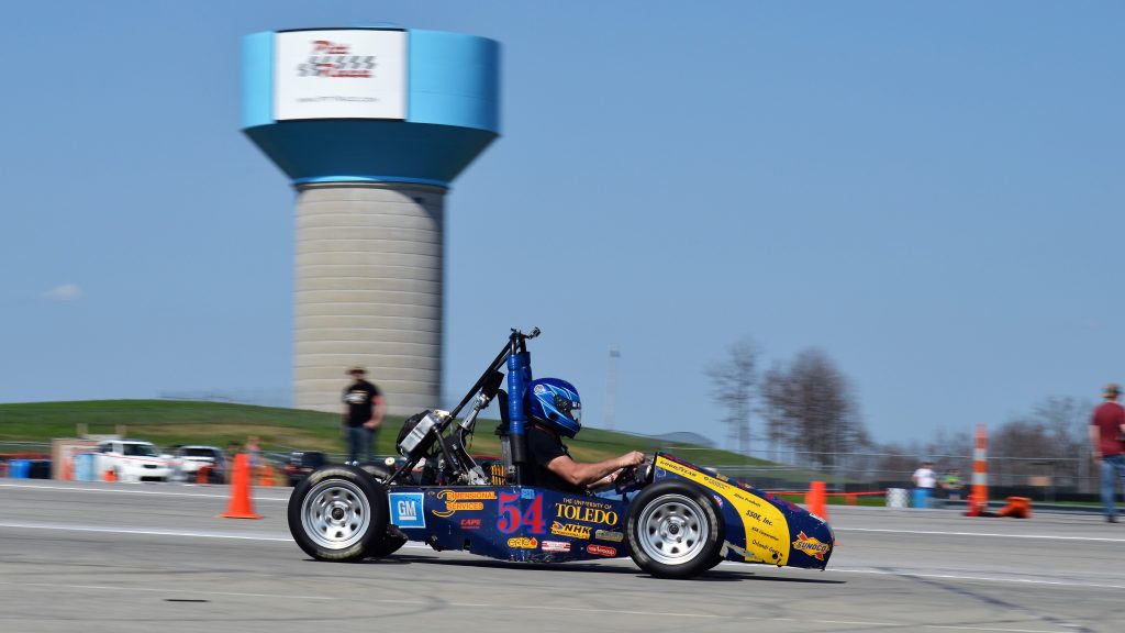 Geoffrey sits in the racing car, the background is blurred. You can see a tower, more cars and people near the race track. His car is blue and yellow and has lots of stickers on it. He himself wears a black T-shirt and a blue helmet.