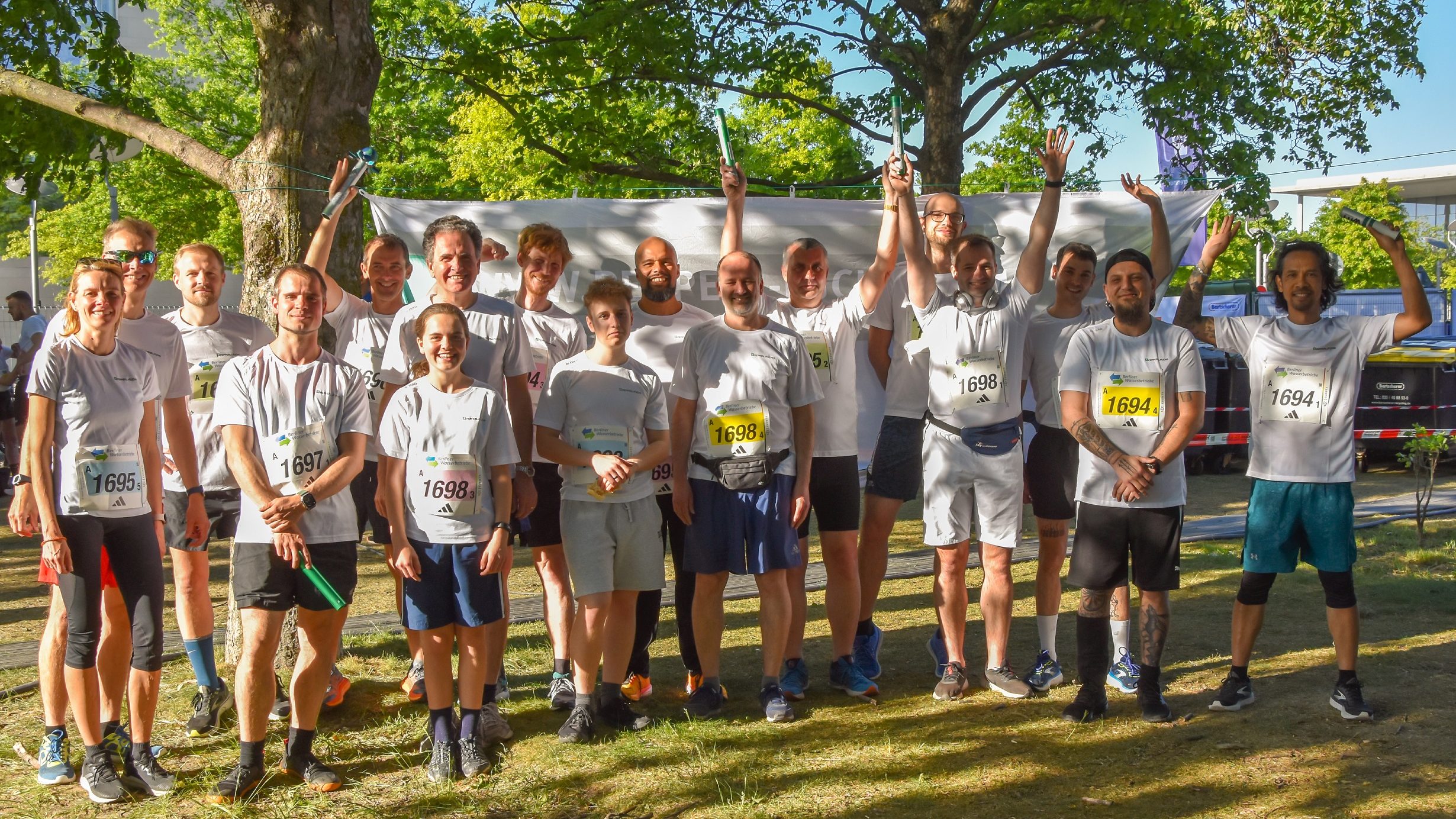 Group photo of the Pepperl+Fuchs runners at the relay race in Berlin. They are standing on a green meadow and some trees can be seen behind them. Three of them raise the relays. They wear the white Pepperl+Fuchs running shirts.