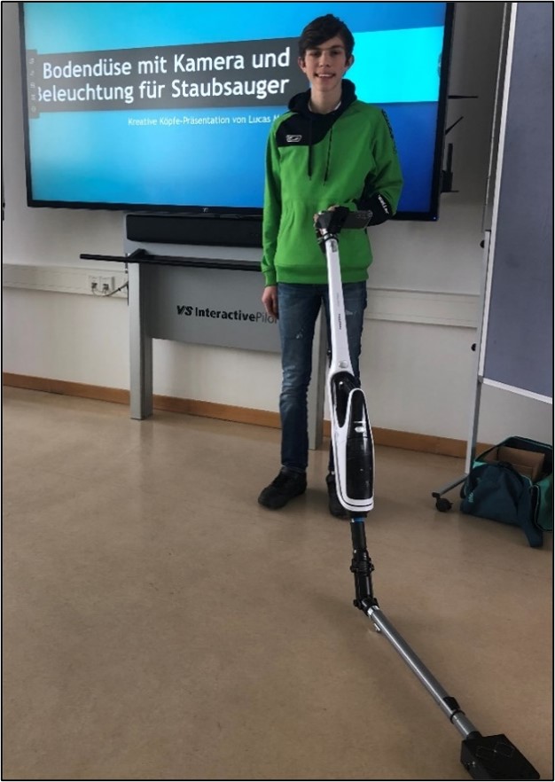 Lucas stands in front of a large television with the finished vacuum cleaner and smiles proudly into the camera. His project title “Floor nozzle with camera and lighting for vacuum cleaners” can be read again on the television.