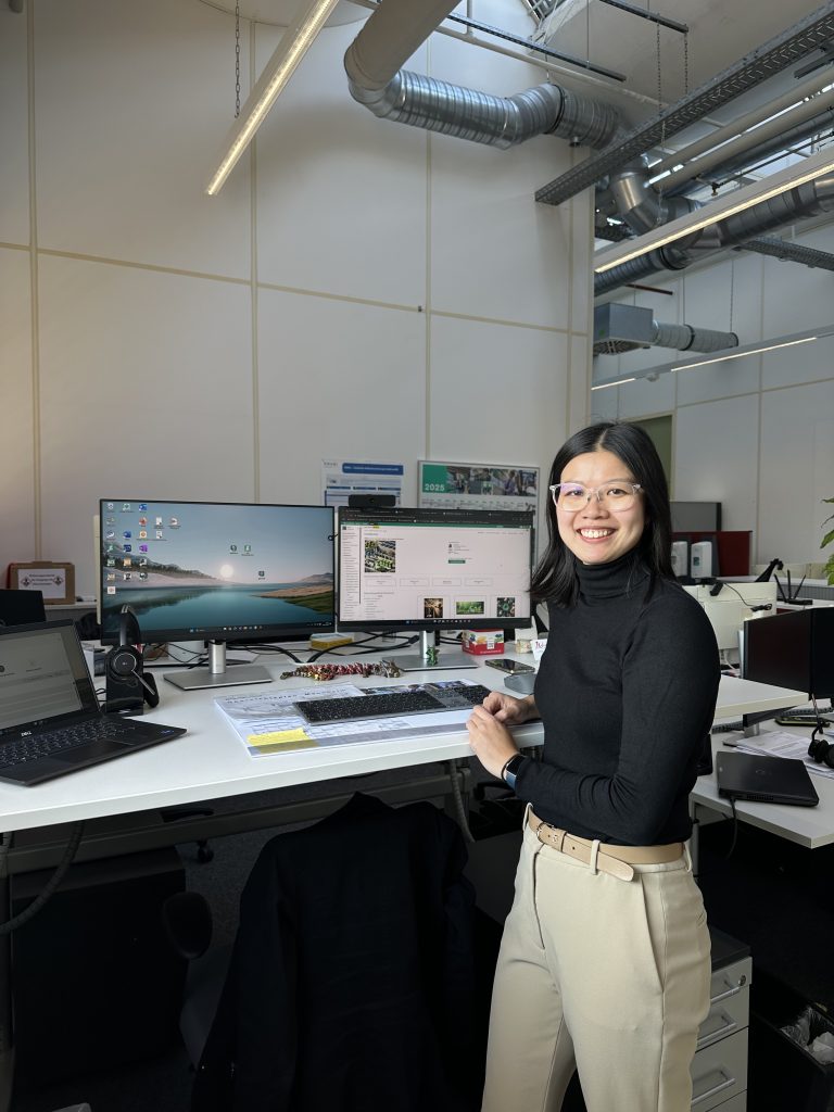 Yun Xia stands at her workstation at Pepperl+Fuchs and looks into the camera. On her desk are a laptop, two monitors, and various office supplies.