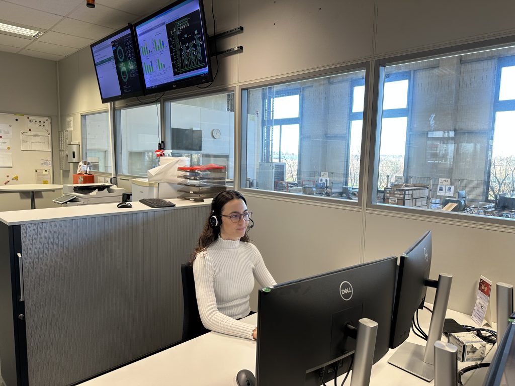 Rebecca is a project and process engineer at Pepperl+Fuchs. Here she is sitting at her desk, working. In the background, you can see other workstations and two monitors hanging on the wall above her.