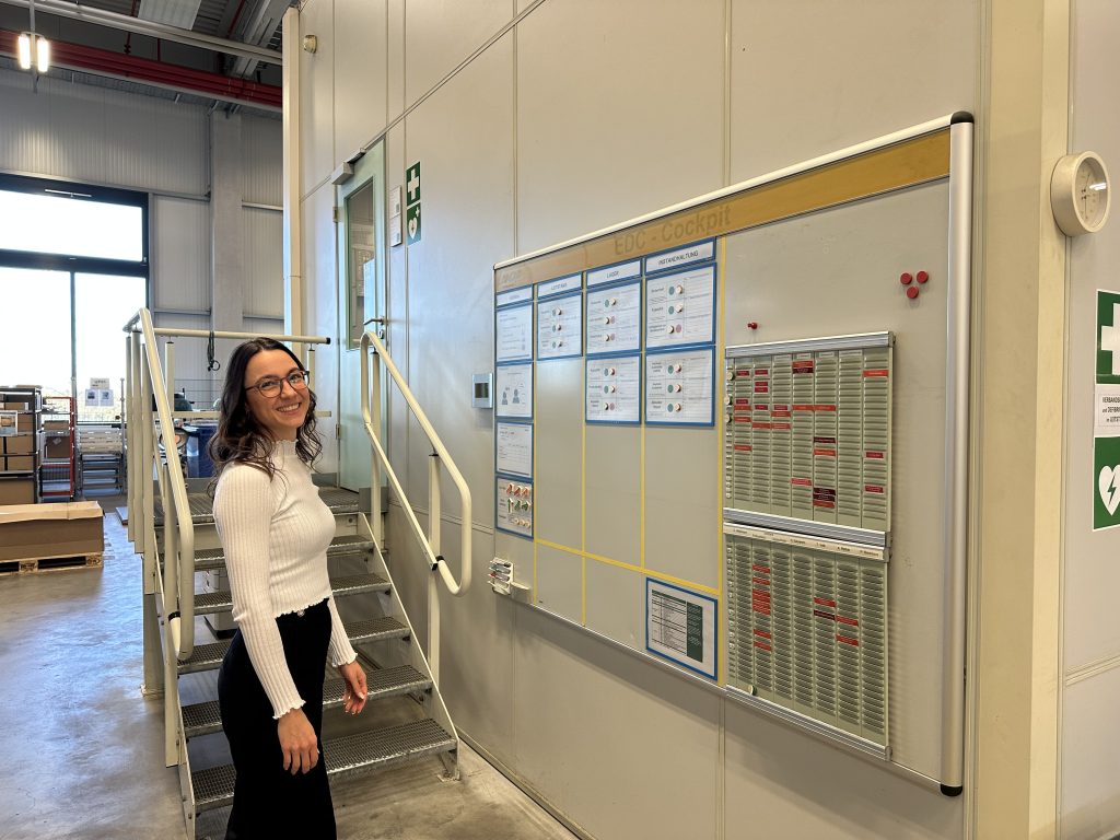 Rebecca walks through Pepperl+Fuchs' logistics department. To her right hangs a large board displaying various information.