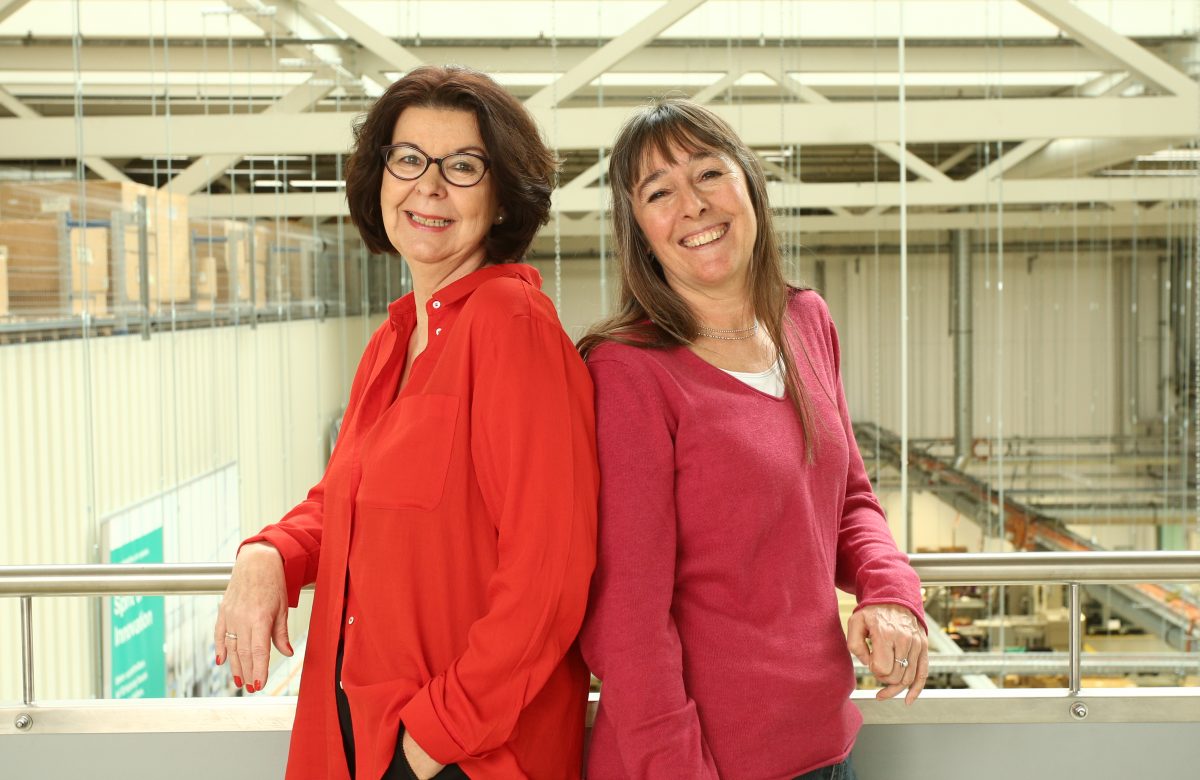 40 Jahre Firmenzugehörigkeit: Claudia und Cosima stehen auf einer Galerie, im Hintergrund sieht man unten die Produktionshalle. Sie strahlen in die Kamera.