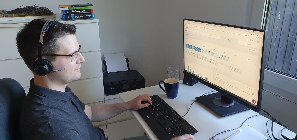 Matthias, Social Media Manager bei Pepperl+Fuchs, an seinem Arbeitsplatz im Home Office. Er trägt ein Headset und schaut auf seinen Bildschirm. Hinter ihm steht eine weiße Kommode mit Fachliteratur darauf.